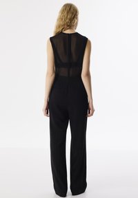Jump suit nero senza maniche con pannello trasparente sul retro, scollatura alta e pantaloni a gamba larga. Linee pulite e design minimalista, presenta chiusura con zip sul retro.