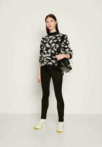 Mulher vestindo um suéter com padrão preto e branco, calças pretas, ténis brancos com solas amarelas, segurando uma bolsa preta no ombro.