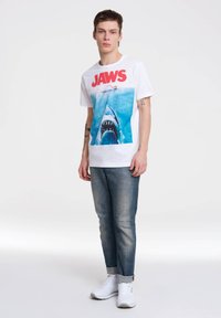 Camiseta de algodón blanca con un gráfico de un tiburón azul y el texto rojo "JAWS". El modelo lleva puestos unos jeans de mezclilla claros y zapatillas blancas.