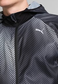 Giacca leggera nera e grigia con strisce orizzontali, cappuccio e chiusura con zip. Presenta un logo bianco nella zona del petto. Tessuto morbido.