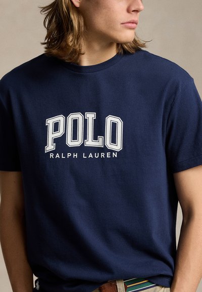 Young person with shoulder-length hair wearing a navy blue Polo Ralph Lauren t-shirt, with head turned to the side.