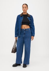 Blue denim jacket with brown collar, black cropped top, wide-leg blue jeans, and black chunky boots, holding a small brown handbag.