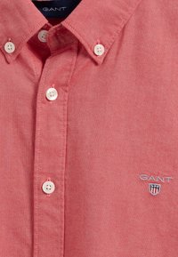 Czerwona koszula na guziki wykonana z fakturowanej tkaniny, z klasycznym kołnierzem, białymi guzikami oraz małym haftowanym logo na piersi.