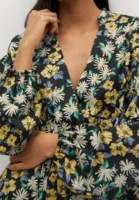 Kukalli wraps-top, joka esittelee mustaa taustaa keltaisilla ja valkoisilla päivänkakkaroilla, puffeilla hihoilla ja syvällä V-kaula-aukolla. Pehmeä kangas, jossa on kevyt kiilto.