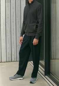 Szary sweter zapinany na zamek z teksturowanym wzorem, w połączeniu z luźnymi granatowymi spodniami i szarymi butami sportowymi, stojący na tle drewnianej ściany.