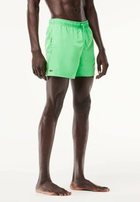 Helle grüne Badeshorts aus leichtem Material mit einem elastischen Bund und Kordelzug sowie seitlichen Taschen. Sichtbares goldenes Markenlogo.