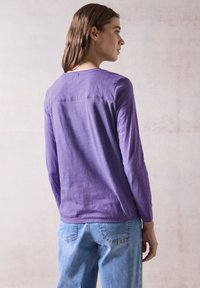 Top lungo viola a maniche lunghe con scollatura rotonda, in tessuto leggero e con un dettaglio cucito sulla parte superiore della schiena, abbinato a jeans azzurri chiari.