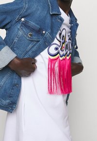 Jeansjack met zilveren knoop, wit shirt met kleurrijke geborduurde letters en roze franjes, dat gelaagde texturen benadrukt.