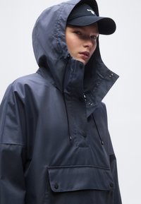 Άτομο που φοράει μπλε σκούρο αδιάβροχο με κουκούλα και μπροστινή τσέπη, καθώς και μαύρο καπέλο μπέιζμπολ, κοιτώντας λίγο πλάγια μπροστά σε απλό φόντο.