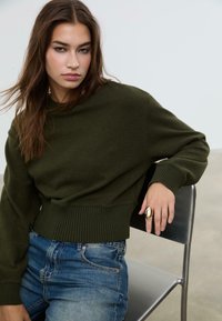 Giovane donna con lunghi capelli castani seduta su una sedia di metallo, indossa un maglione verde oliva, jeans blu e un grande anello d'oro sulla mano destra.