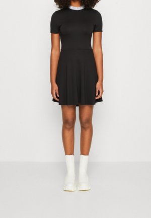 Femme portant une robe noire à manches courtes avec une jupe évasée et des bottines blanches, debout devant un fond clair uni.