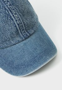 Cappellino in denim con visiera curva, colore blu scuro, cuciture visibili e superficie texturizzata. Al primo piano si vedono i dettagli della tessitura del tessuto.