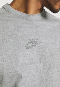 Pelēka kokvilnas T-kreklu ar teksturētu, mirdzošu Nike logotipu uz krūtīm. Iezīmē klasisku apļveida kakla dizainu un īsām piedurknēm.