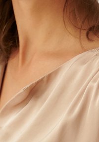Tessuto beige chiaro con morbide drappeggiature, caratterizzato da una texture liscia e un collo a V poco profondo. Pieghe sottili valorizzano la zona delle spalle.