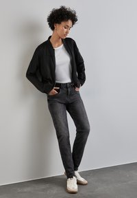 Chaqueta bomber negra sobre una camiseta blanca, combinada con jeans oscuros y desgastados y zapatillas blancas. Cabello rizado enmarca el rostro. Fondo minimalista.