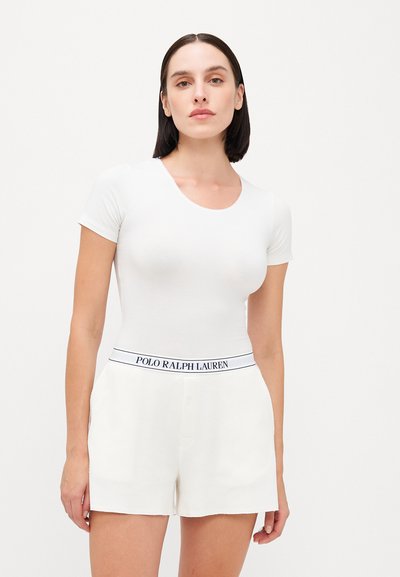 Fehér, rövid ujjú, testhezálló felső, krémszínű rövidnadrággal párosítva, amelyen kék "POLO RALPH LAUREN" derékpánt és gomb részlet található. Sima anyagú textúra.