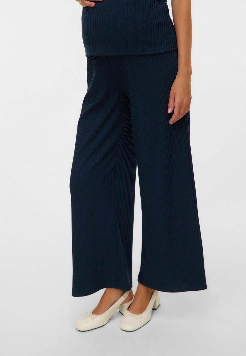 Pantalon large côtelé bleu marine avec une texture lisse, associé à des chaussures de couleur claire ornées d'un nœud. Design simple et décontracté.