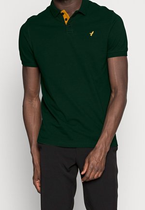 Man draagt een donkergroen poloshirt met korte mouwen, een gele knopenbies en een klein geel vogel-logo op de borst, gecombineerd met een zwarte broek.