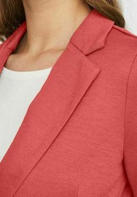 Punainen tekstuuri-taskutakki, jossa on viisto kaulus, esillä valkoisen paidan päällä. Kangas on kudottu, ja istuvuus näyttää olevan muotoiltu.