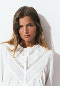 Blusa blanca con un tejido texturizado, que presenta un cuerpo de encaje y un diseño plisado. Botones en la parte delantera y mangas suaves y fluidas.