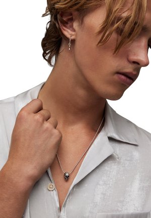 Silver skull pendant necklace on a beaded chain, worn over a light gray, textured button-up shirt. Hair styled in loose waves.
