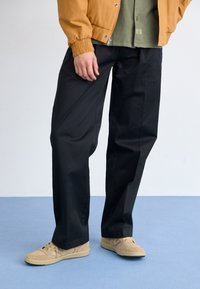 Pantaloni neri a gamba larga abbinati a una camicia verde e una giacca bomber senape. Scarpe da ginnastica beige completano l'outfit. Texture del tessuto liscia evidente.