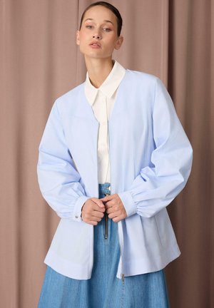 Jeune femme portant une veste zippée bleu clair par-dessus une chemise blanche à col et une jupe bleue, debout devant un fond de rideau beige.