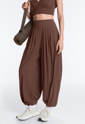 Žena v hnedých plisovaných širokých nohaviciach a crop tope, drží zvinutú jogovú podložku, v kombinácii s bielymi športovými topánkami.