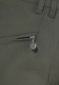 Primer plano de tela verde oscura con un bolsillo con cremallera parcialmente abierto y tirador redondo de cremallera plateado con patrón texturizado.