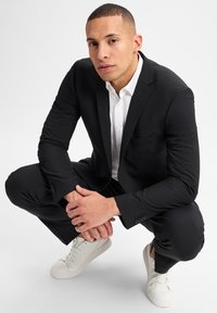 Zwart pak met een getailleerd ontwerp, witte overhemdd met een textuurstof. Sneakers in crèmekleur. Onderwerp in hurkpositie met handen samengevouwen.