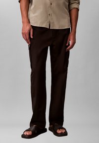 Homme portant un pantalon cargo marron foncé et des sandales noires, debout devant un fond gris uni, les mains détendues le long du corps.