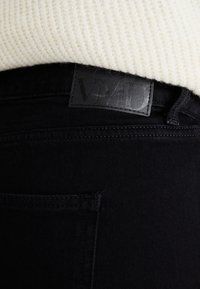 Schwarze Denim-Jeans mit einem mattschwarzen Lederetikett, das einen geprägten Text aufweist. Der Stoff wirkt glatt mit einer engen Webart.