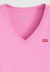 Różowa bawełniana koszulka z dekoltem w serek z czerwoną naszywką logo Levi's na piersi, przedstawiająca szczegółowy dekolt i strukturę materiału.