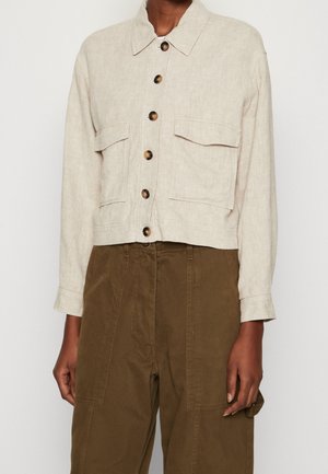 Veste beige boutonnée avec grandes poches avant, associée à un pantalon marron taille haute avec des détails de coutures apparentes.