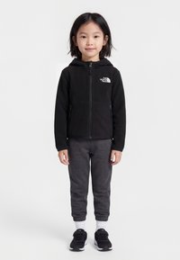 Μικρό παιδί όρθιο, φορώντας μαύρο φούτερ με κουκούλα και φερμουάρ με λογότυπο, σκούρο γκρι φόρμα, λευκές κάλτσες και μαύρα αθλητικά παπούτσια.