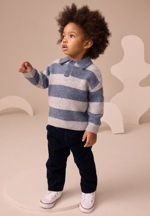 Dieťa s kučeravými vlasmi v modro-šedom pruhovanom svetri, námorníckych nohaviciach a bielych teniskách, stojace na okrúhlom podstavci.