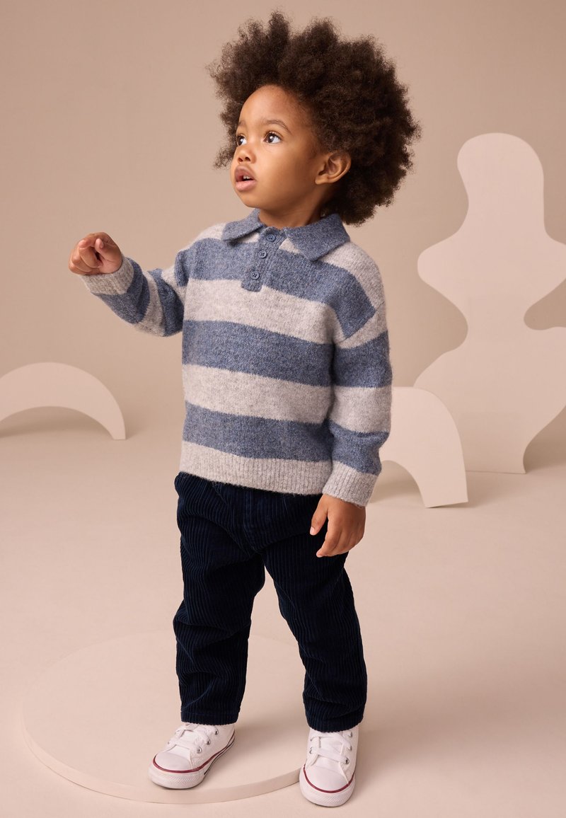 Malček z kodrastimi lasmi, oblečen v modro-sivo črtast pulover, temno modre hlače in bele superge, stoji na okroglem odru.
