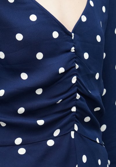 Φόρεμα σε ναυτικό μπλε με λευκές πουά, με βαθύ V λαιμόκοψη και ρυτιδωτό σχέδιο στο μπούστο. Λείο, ελαφρύ ύφασμα.