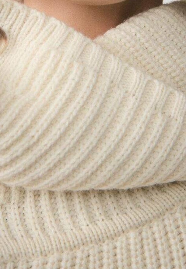 Gros plan d'une personne portant un pull en maille côtelée de couleur crème, montrant la texture et les détails du tissu.