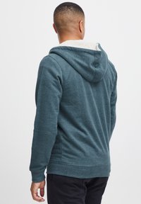 Mikina s kapucí v teplém teal odstínu s měkkou, béžovou podšívkou. Disponuje dlouhými rukávy a žebrovaným lemem. Pohled zezadu, ukazující celkový design.