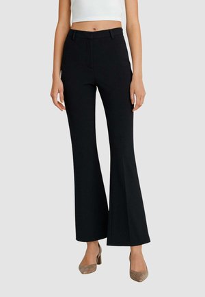 Pantaloni neri a palazzo realizzati con un materiale elasticizzato, caratterizzati da una vita aderente, passanti per la cintura e una finitura liscia. Abbinati a tacchi beige.