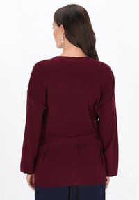 Bordowy sweter dzianinowy z długimi rękawami, prążkowaną teksturą i wiązanym paskiem w talii. Detale przy ramionach z ozdobami.