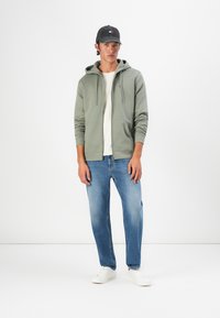 Sudadera con capucha y cremallera en un verde oliva, combinada con una camiseta blanca y jeans de un azul claro. Accesorios incluyen una gorra oscura y zapatillas blancas.