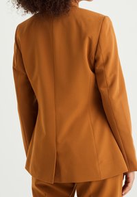 Brun skräddarsydd kavaj med en smal passform, vassa slag och ventilerad rygg. Tillverkad av ett mjukt tyg med en subtil lyster, kombinerad med matchande byxor.