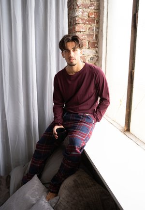 Donkerrood shirt met lange mouwen, gecombineerd met marineblauwe en rode geruite pyjamabroek. Zittend op een vensterbank met een zwart kopje, omgeven door kussens.