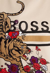 Le tissu beige présente un tigre brodé en brun et noir avec des accents floraux rouges, violets et blancs ainsi que des lettres audacieuses "BOSS".