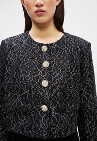 Giacca nera corta con ricami floreali argento, dotata di tre bottoni decorativi in cristallo e un colletto rotondo. Tessuto testurizzato con tasche.