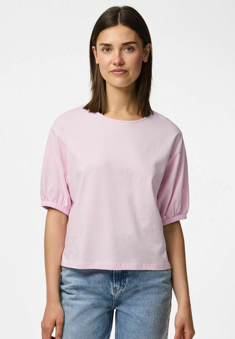 Vaaleanpunainen lyhythihainen croppit-paita, jossa on puffy hihat ja pyöreä kaula-aukko. Valmistettu pehmeästä puuvillakankaasta. Rennon mallinen. Vaaleansiniset farkut.