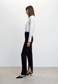 Schwarze maßgeschneiderte Hose mit seitlichen Schlitzen, kombiniert mit einem weißen Langarmoberteil. Das Model steht seitlich auf einem hellgrauen Boden vor einer schlichten Wand.