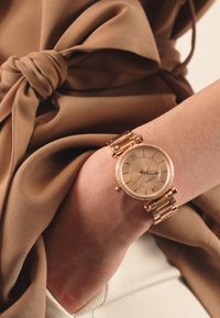 Montre-bracelet en or avec un cadran rond et une lunette texturée. Le bracelet est composé de maillons entrelacés. Portée sur un poignet couvert par un blouse beige.